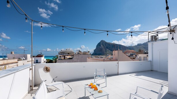 A rooftop terrace with white chairs, a table with drinks, and string lights. A mountain and sea are visible in the background.