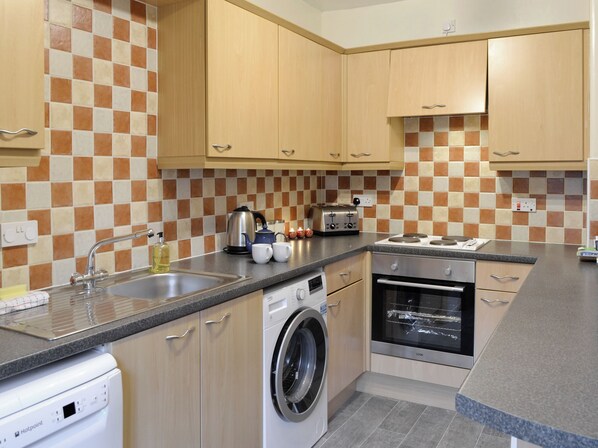 A modern kitchen with wooden cabinets, a washing machine, a dishwasher, and a built-in oven. - Baldowrie Farm Cottage (Kettins, near Blairgowrie)