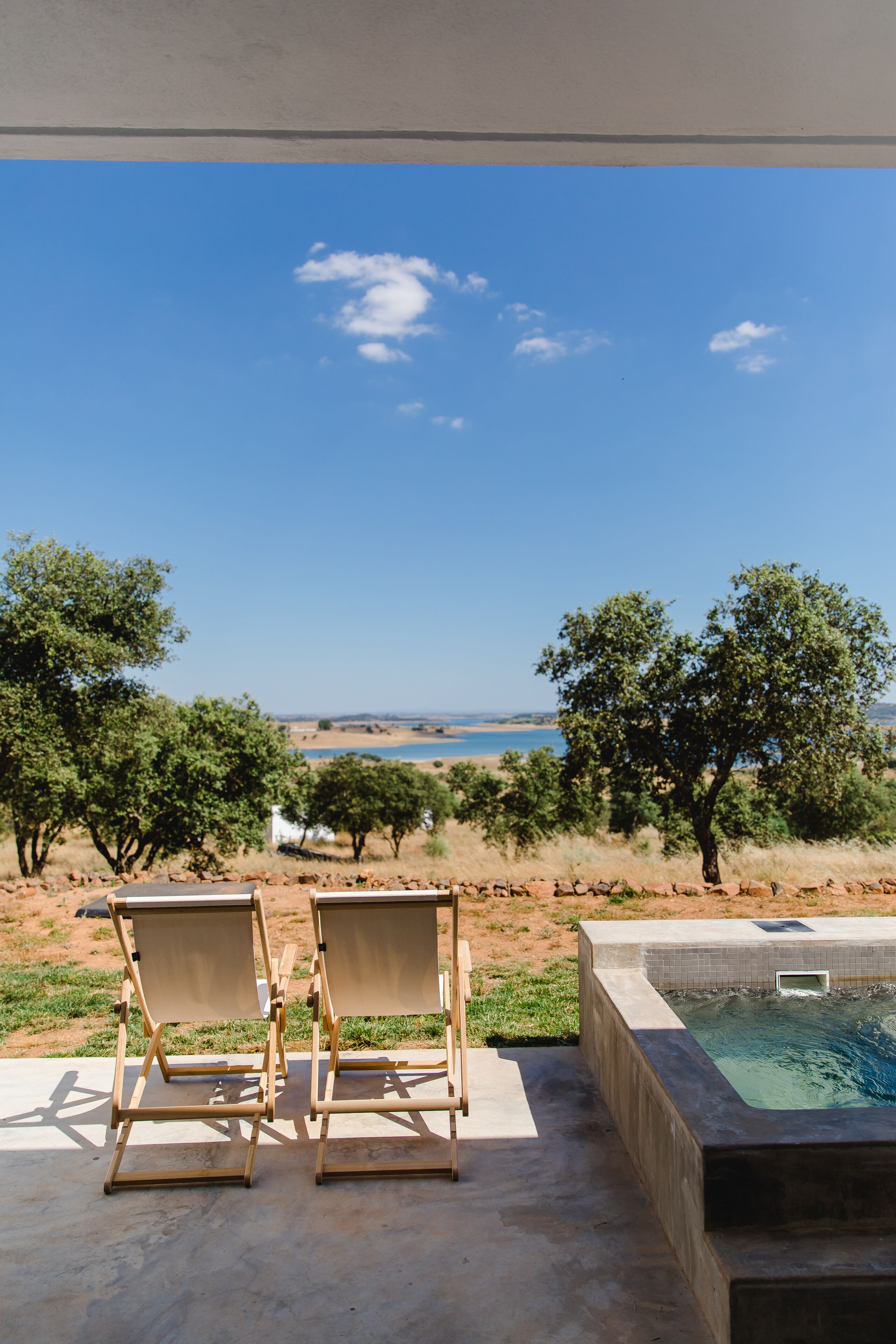A pool area with two lounge chairs facing a scenic view of a body of water and trees.