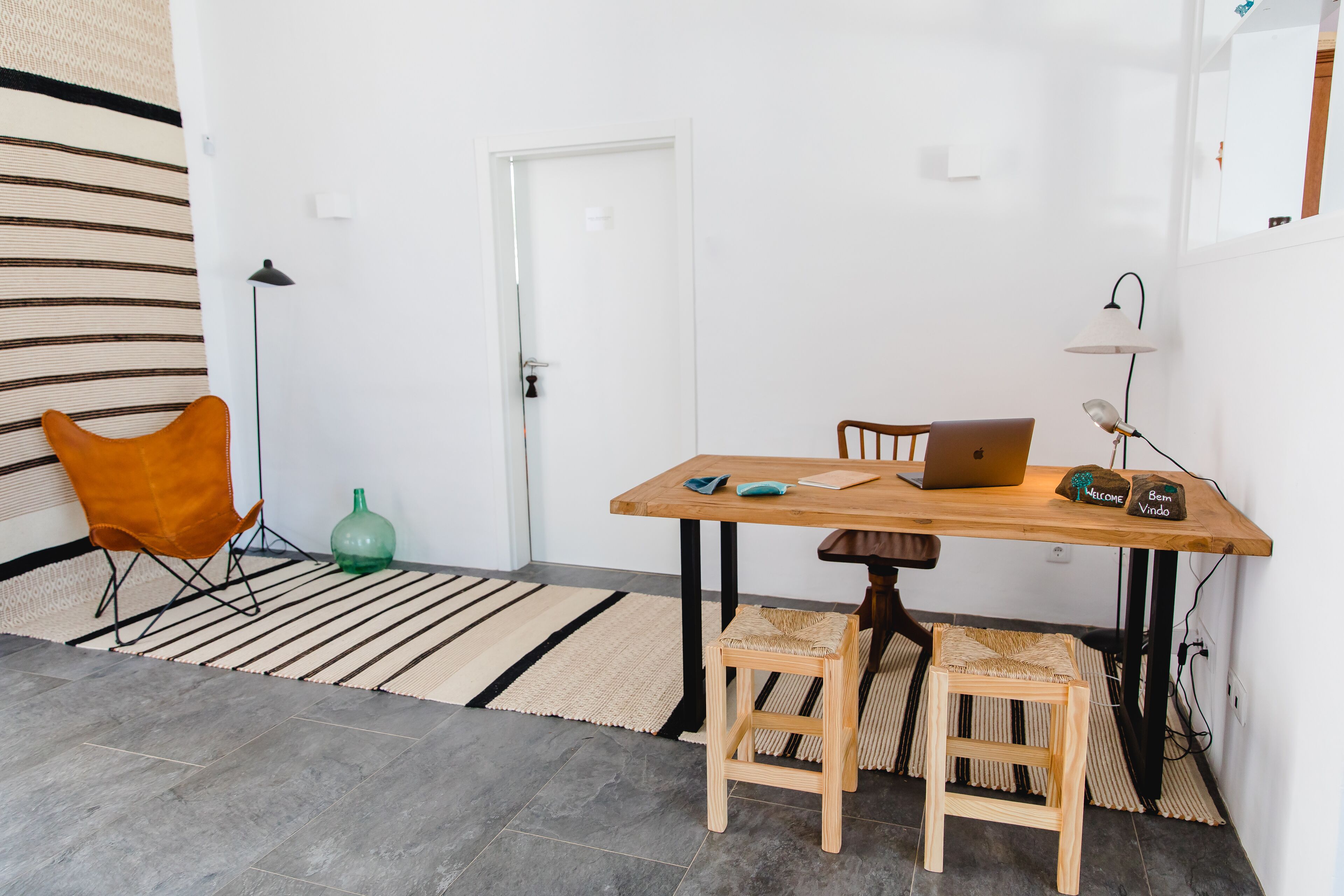 A room with a wooden table, two chairs, a laptop, a green bottle, and a striped rug.