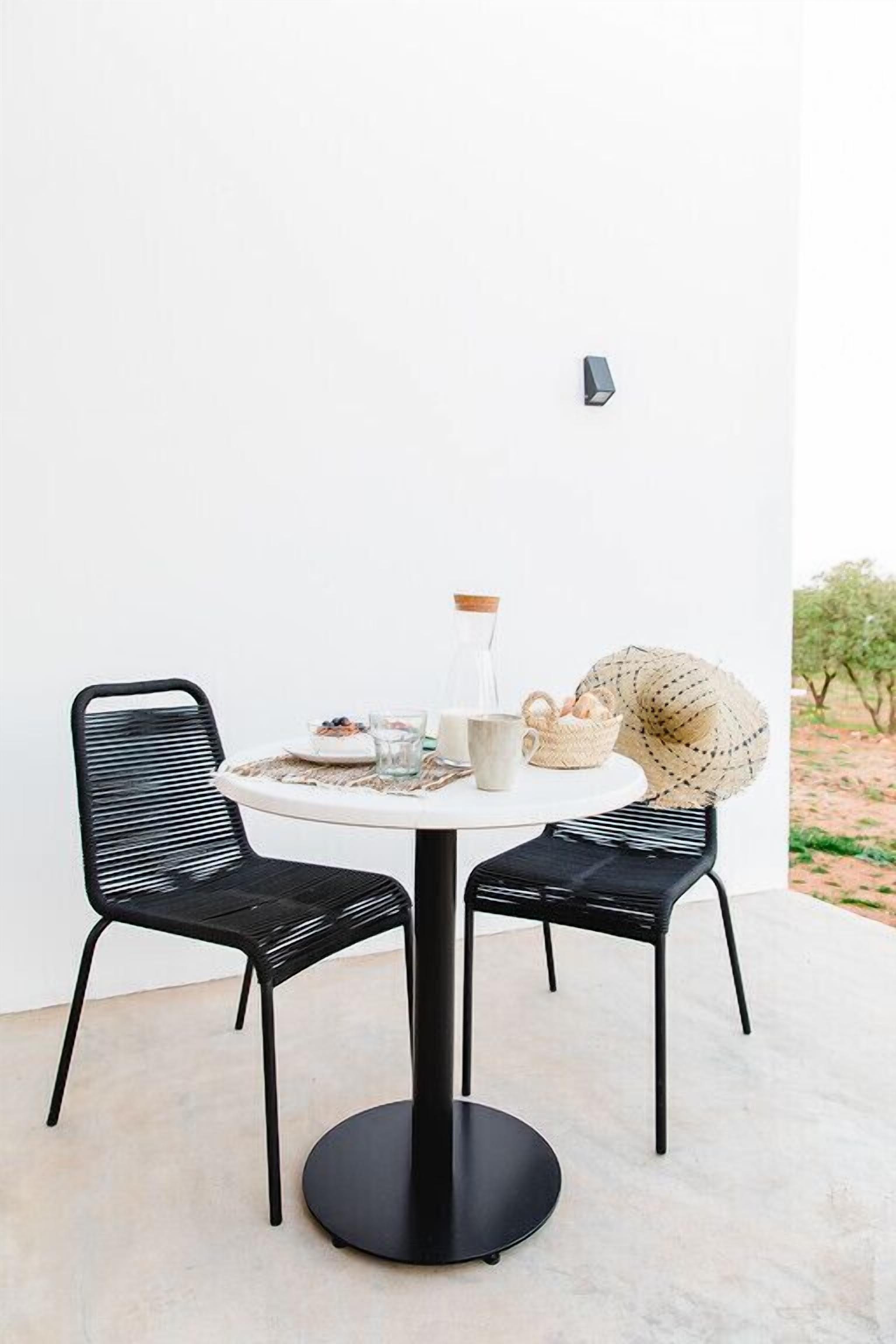 Una mesa redonda con dos sillas negras, una pared blanca y un pequeño espacio exterior con vista a árboles.
