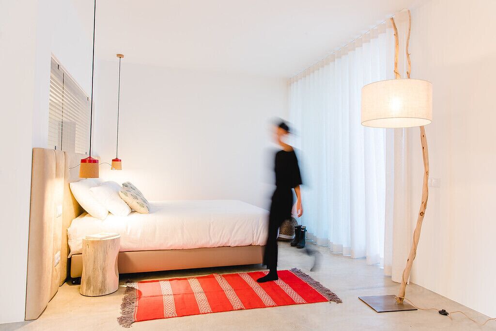 Un dormitorio moderno con una cama, un tapete a rayas rojo y blanco, una mesita de noche de madera y una lámpara de pie.