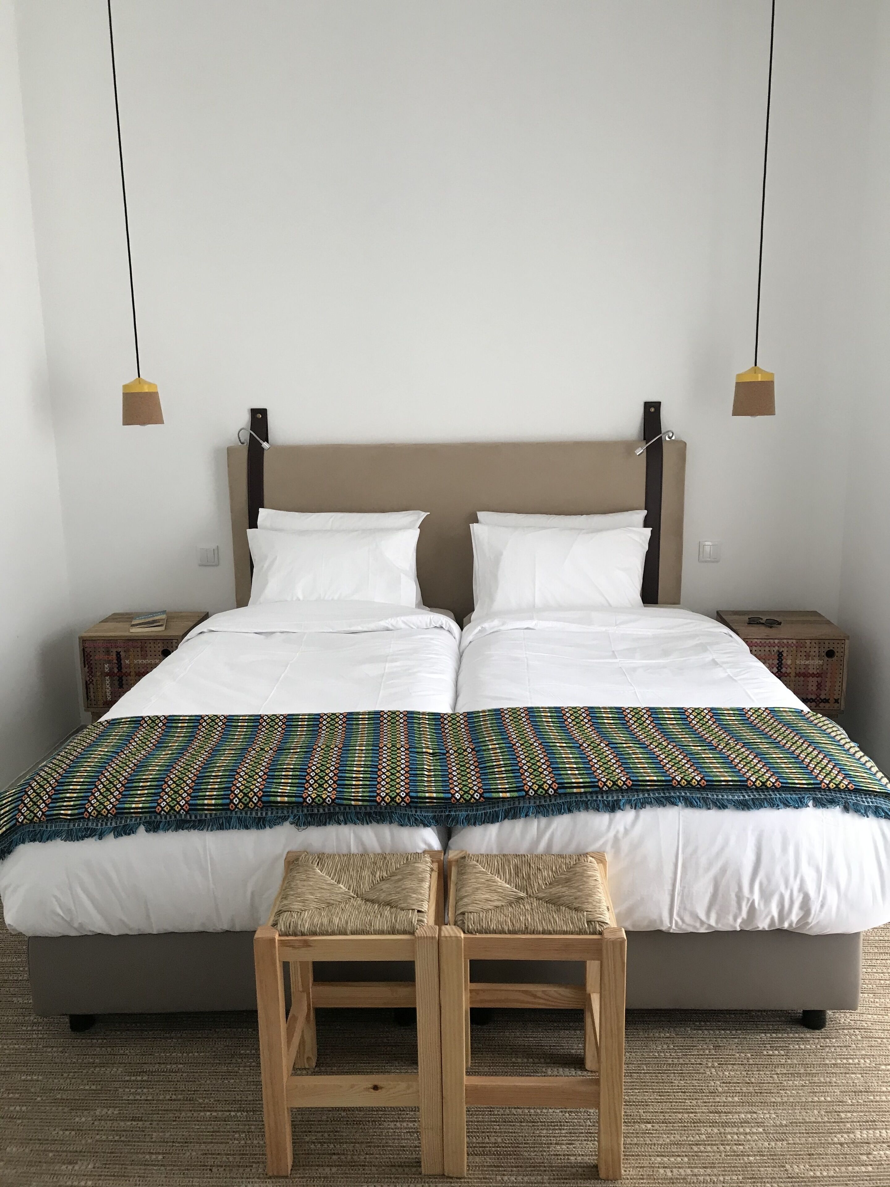 Uma cama de casal com lençóis brancos, um cobertor colorido estampado e duas cadeiras de madeira na frente.