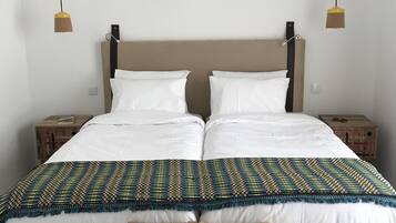 Uma cama de casal com lençóis brancos, um cobertor colorido estampado e duas cadeiras de madeira na frente.