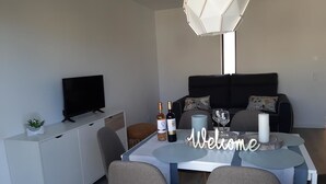 Een moderne woonkamer met een eettafel, twee grijze stoelen, een witte kast, een flatscreen televisie, een bank met kussens, een decoratief bord met de tekst 'Welcome' en een hanglamp.