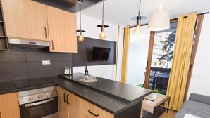 A modern kitchen with wooden cabinets, a black countertop, and stainless steel appliances. There is a flat-screen TV mounted above the counter and a small table with a plant.