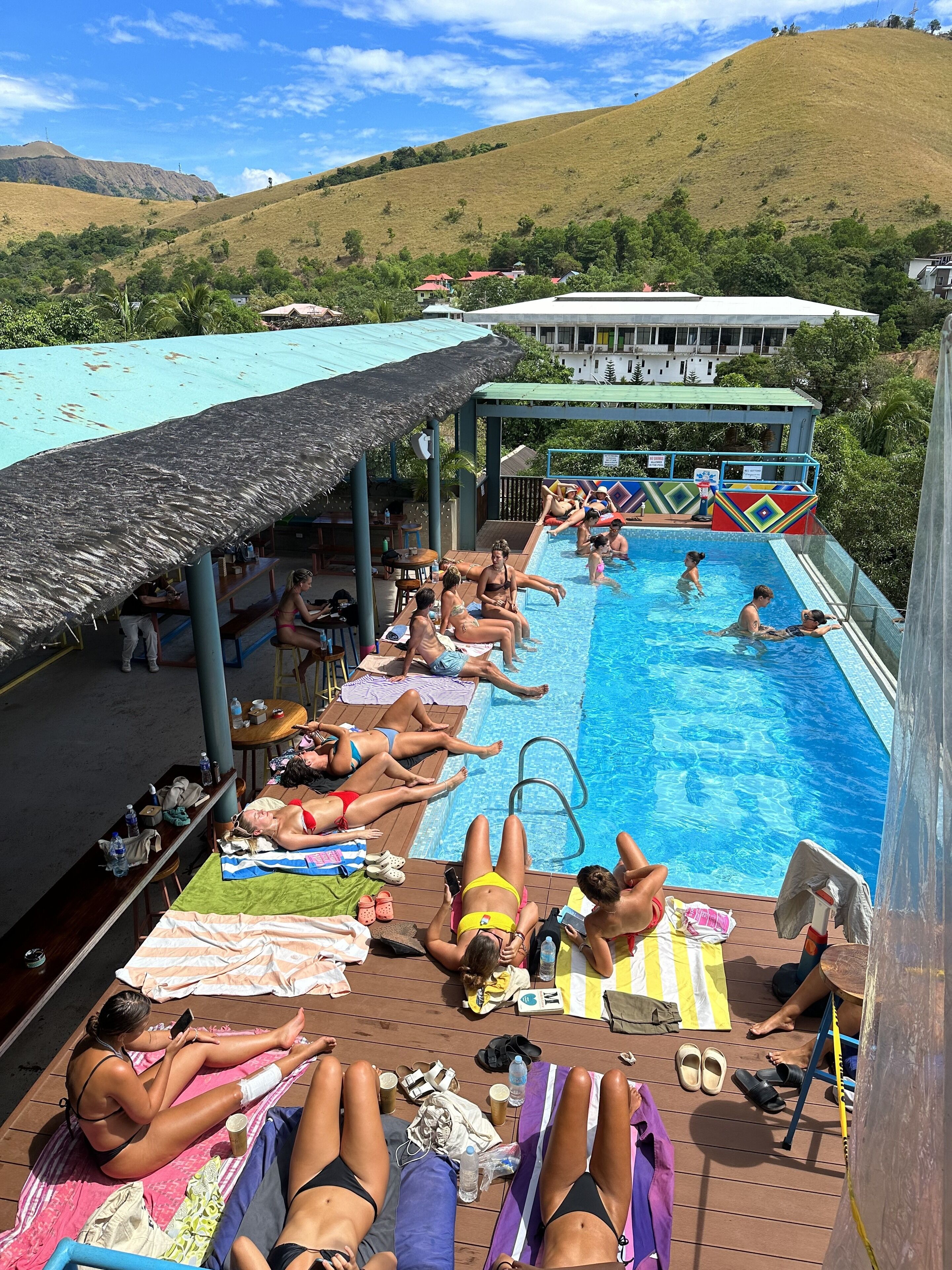 Une piscine entourée de personnes qui prennent des bains de soleil et se baignent, le tout abrité par une structure à toit de chaume, avec une vue sur des collines en arrière-plan.