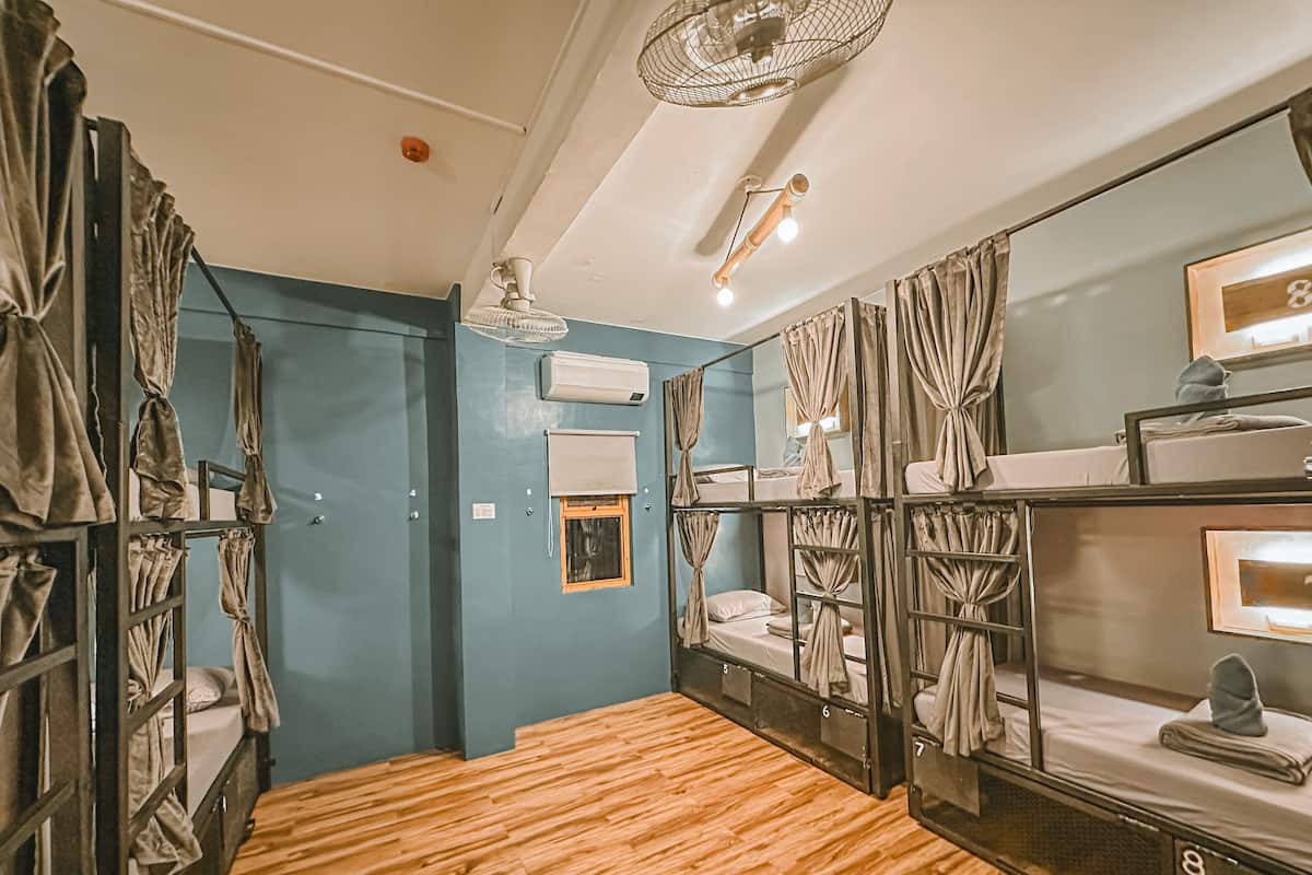 A dormitory room with bunk beds, a wooden floor, a ceiling fan, and a small window with curtains.