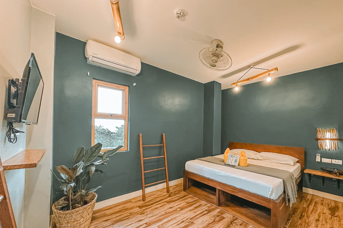 A bedroom with a bed, wooden floor, a window, a wall-mounted TV, an air conditioner, a ceiling fan, and a potted plant.