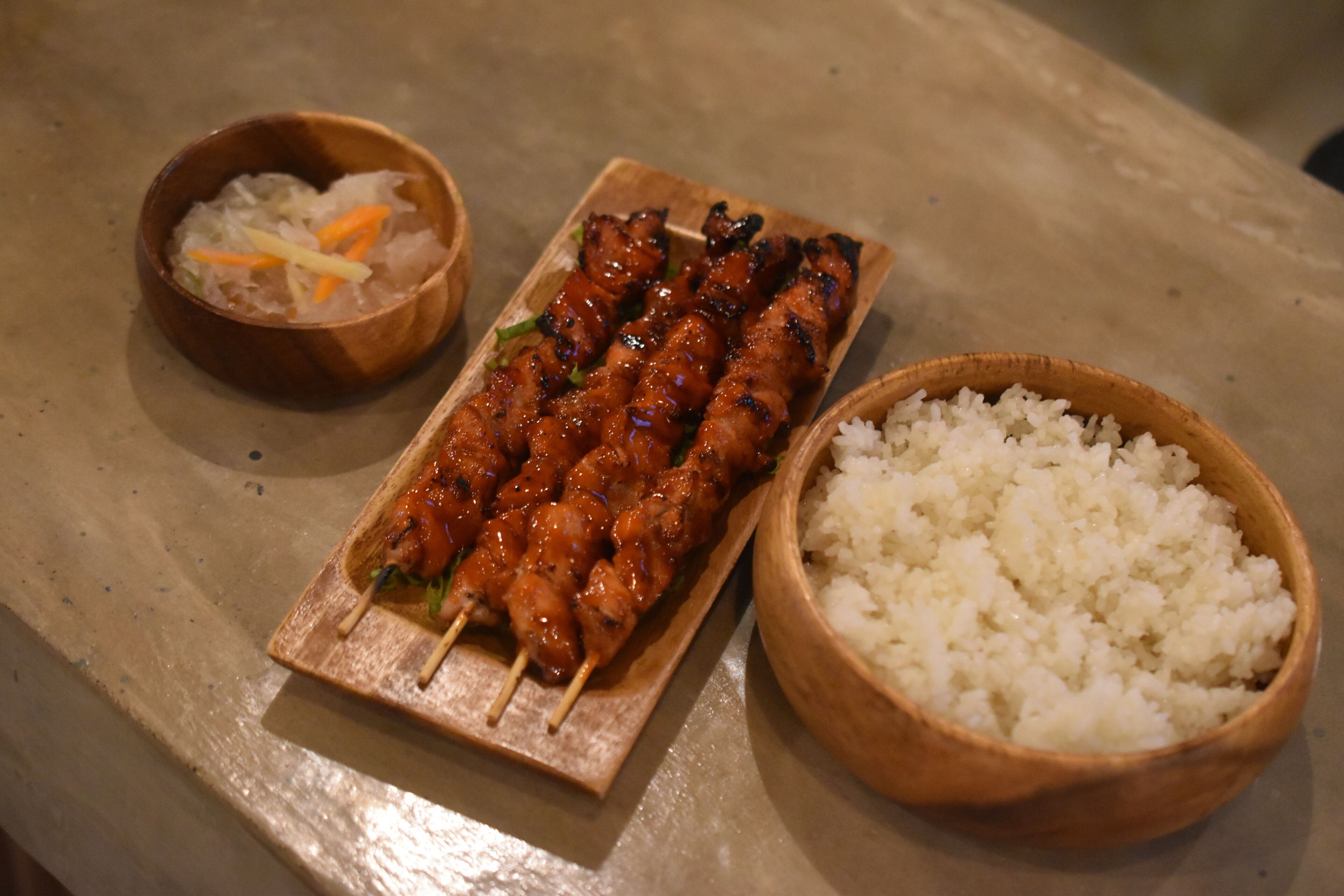 Una bandeja de madera con brochetas de carne a la parrilla, un tazón de arroz blanco y un recipiente pequeño con verduras encurtidas.
