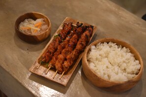 A wooden tray with skewers of grilled meat, a bowl of white rice, and a small bowl of pickled vegetables. - Outpost Hostel - Coron - Adults Only (Coron)