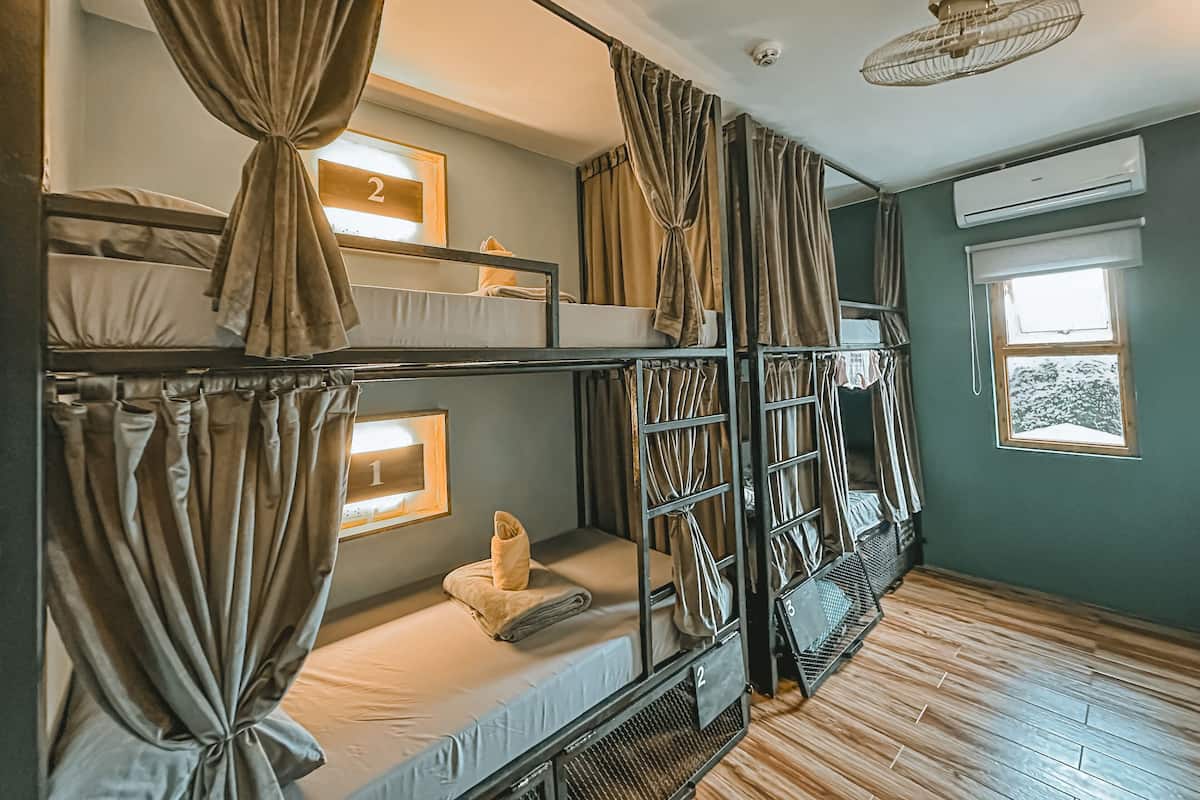 A dormitory room with bunk beds, curtains, a ceiling fan, an air conditioning unit, and a window with a view of trees.
