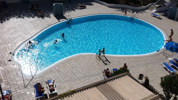 Ein Schwimmbecken mit Fliesen umrandet, Liegestühlen und einigen darin schwimmenden Personen.