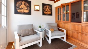 A room with two white armchairs, a small side table with a vase and books, and a wooden cabinet with glass doors. - Jacuzzi Garden Holiday Home (Playa del Inglés)