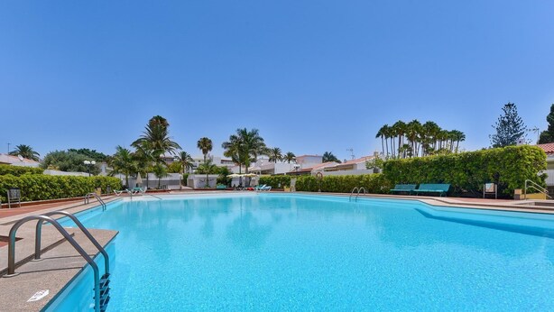 A clear blue swimming pool with a metal ladder, surrounded by palm trees and a row of beach chairs. - Jacuzzi Garden Holiday Home (Playa del Inglés)