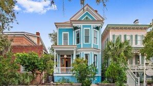Un edificio de estilo victoriano con ventanal saliente, un porche y un pequeño jardín con árboles y arbustos.