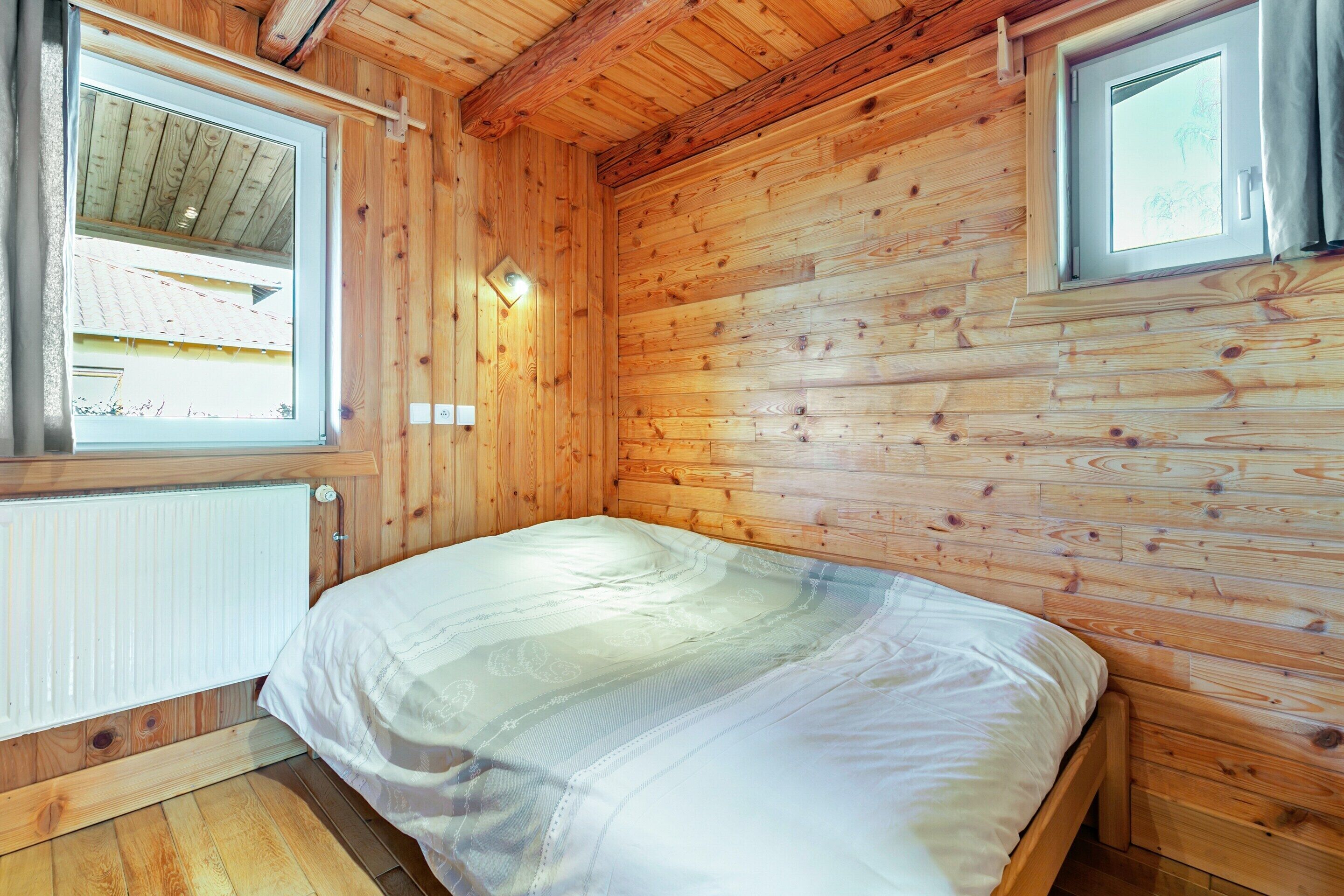 A wooden cabin room with a bed, two windows, and a radiator.