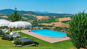 Une piscine entourée de chaises longues et de parasols, bordée d’une pelouse et offrant une vue panoramique sur un paysage pittoresque composé de champs et de montagnes.