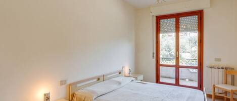 A bedroom with a bed, bedside table, lamp, and a window with a view of greenery.