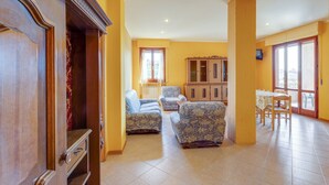 A spacious living area with a wooden cabinet, two patterned armchairs, a dining table with chairs, and a view through large windows. - Apartment in Tuscany Near Lake Trasimeno (Tuoro sul Trasimeno)