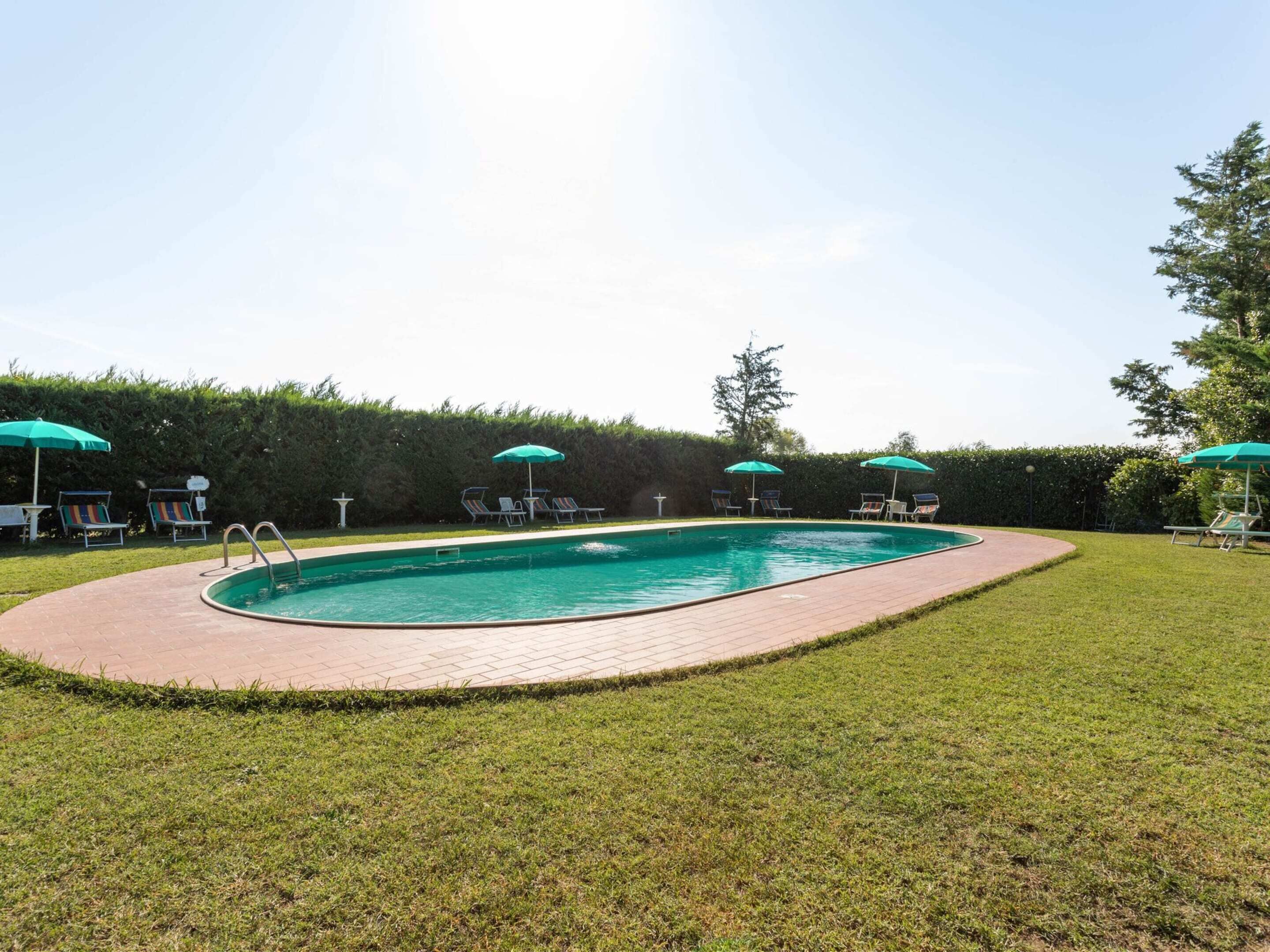 Ein rechteckiger Pool mit Backsteinrand, umgeben von einem gepflegten Rasen und mehreren Liegestühlen mit grünen Sonnenschirmen.
