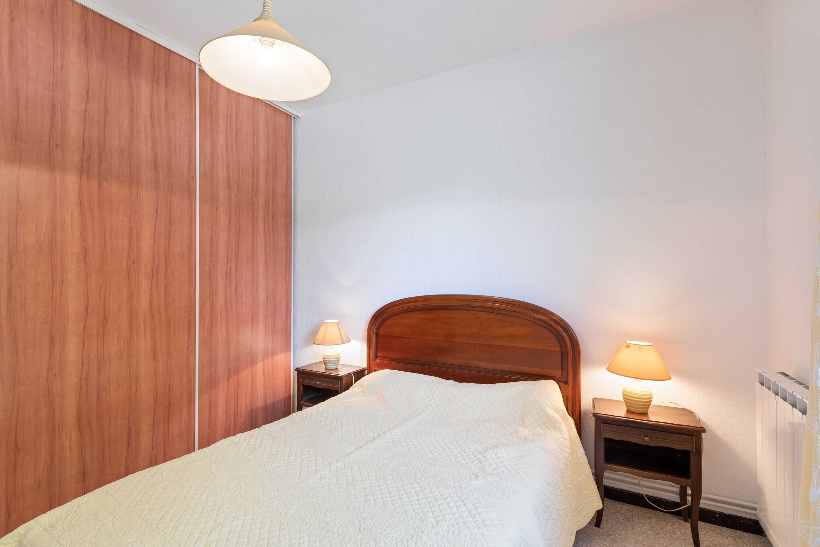 A bedroom with a wooden wardrobe, a bed with a white quilt, two bedside tables with lamps, and a ceiling light.