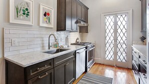 Una cocina moderna con armarios oscuros, azulejos blancos tipo metro, un lavamanos de acero inoxidable y una estufa a gas. Hay dos láminas enmarcadas con motivos botánicos en la pared.