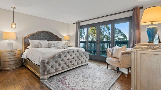 A bedroom with a tufted headboard bed, a large window with a view, a chaise lounge, and a classic lamp.
