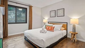 A modern hotel room with a large bed, two bedside lamps, a window with curtains, and two framed artworks on the wall.