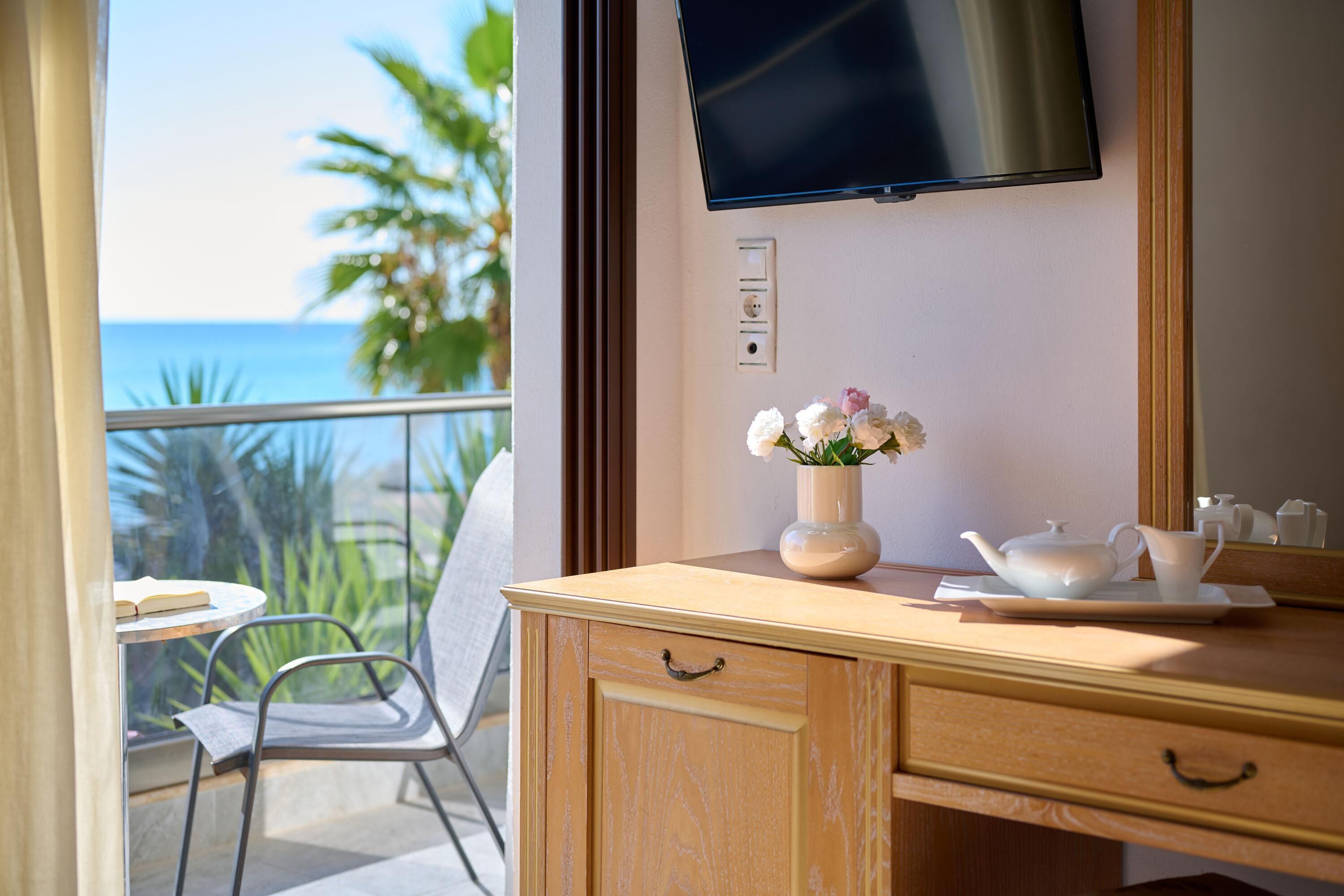 A balcony with a view of the ocean, a chair, a table with a book, a wooden dresser with a vase of flowers, and a flat-screen TV mounted on the wall.