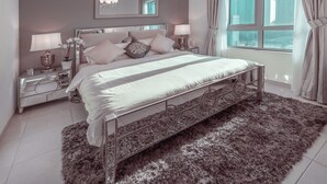 Un dormitorio moderno con una cama grande, mesitas de noche, un espejo y vistas a edificios altos a través de una ventana.