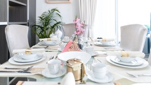 Una mesa de comedor preparada para seis personas, con platos, tazas y cubiertos blancos, un florero con flores rosas y un adorno dorado.