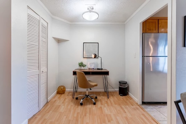 A compact kitchen with a stainless steel refrigerator, a wooden cabinet, and a small workspace with a chair and desk.