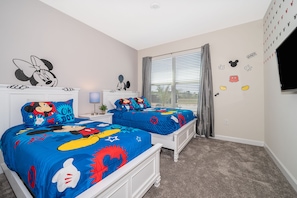 A bedroom with two beds, each covered with a Mickey Mouse themed bedspread. There are wall decals of Mickey Mouse and Minnie Mouse, and a window with blinds.