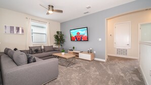 Un salon moderne avec un canapé d’angle gris, une table basse, un téléviseur fixé au mur et un ventilateur de plafond.