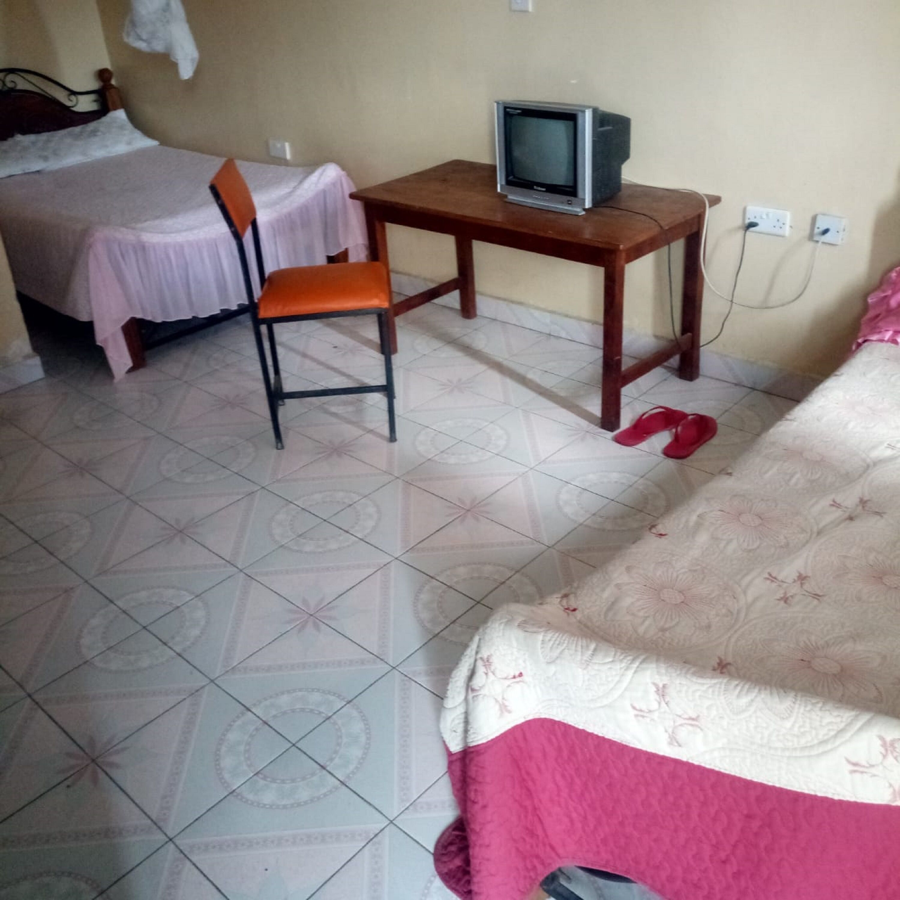 Ein Zimmer mit einem Bett, einem Schreibtisch mit Fernseher, einem Sessel und einem Paar Hausschuhen auf dem Boden.