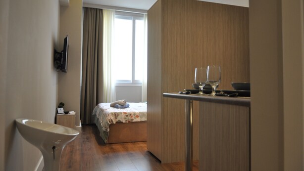 A modern hotel room with a wooden floor, a bed with a floral bedspread, a small bedside table, a TV mounted on the wall, and a window with curtains.