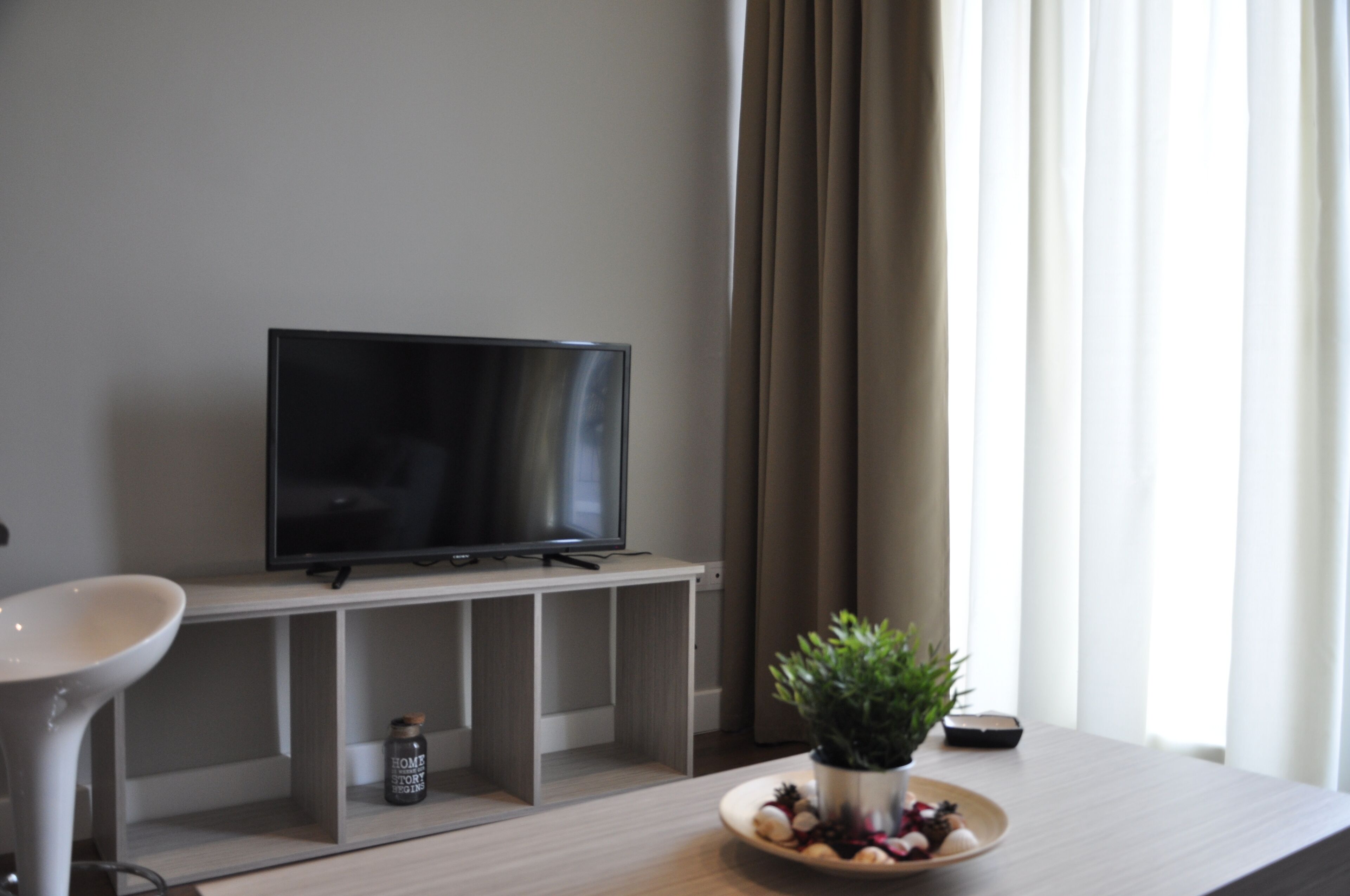 A modern living room with a flat-screen TV on a wooden stand, a white chair, a small table with a potted plant, and a bowl of fruit.