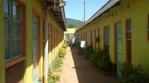 A narrow corridor with yellow walls and green doors, leading to a building with a tiled roof. - Njiwa Guest House (Nakuru)
