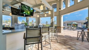 Un vaste espace bar extérieur avec vue sur la piscine, un téléviseur fixé au plafond, ainsi que plusieurs chaises et tables.
