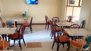 Uma sala de jantar com várias mesas e cadeiras, uma televisão na parede e piso de azulejos.