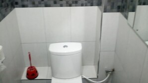 Uma casa de banho pequena com uma sanita branca e um desentupidor vermelho num canto, com paredes em azulejo e chão escuro.