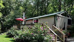 A rustic cabin with a wooden staircase, surrounded by lush greenery and flowering bushes.