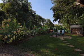 Un jardín con mesa y sillas de picnic, rodeado de árboles y un cobertizo con leña apilada.