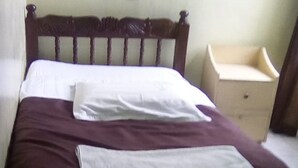 A bed with a wooden headboard, a white pillow, and a purple blanket. There is a small bedside table with a drawer and a nightstand with a lamp. - Pekers Lodge (Nakuru)