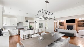 A modern kitchen with white cabinetry, a dining area with a wooden table and white chairs, and a living room with a sofa and a flat-screen TV.