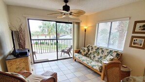 A room with a sofa, two wicker chairs, a glass table, a ceiling fan, a lamp, and a view of the outdoors through a sliding door. - Ocean Village Club C36 (St. Augustine)