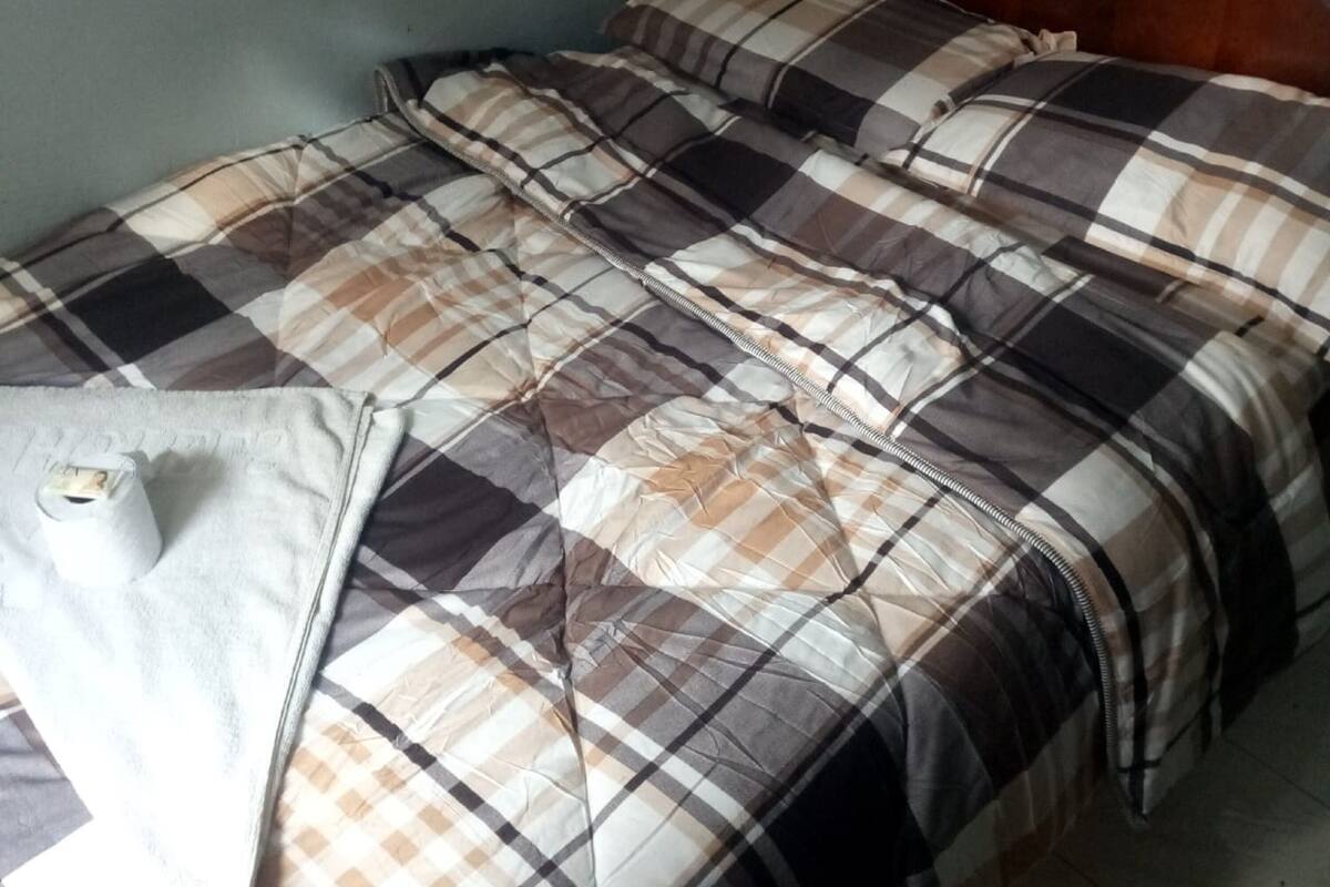 A bed with a plaid bedspread and pillows, a wooden headboard, and a small white object on the left side.
