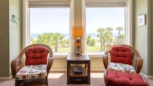A room with two wicker chairs with red cushions, a small table with a lamp, and a view of the ocean through large windows. - Shipwatch #1365 - Heated Pool! (Fernandina Beach)