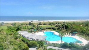 A pool area with a curved design, surrounded by lounge chairs and umbrellas, with a view of the beach and ocean in the background. - Shipwatch #1365 - Heated Pool! (Fernandina Beach)